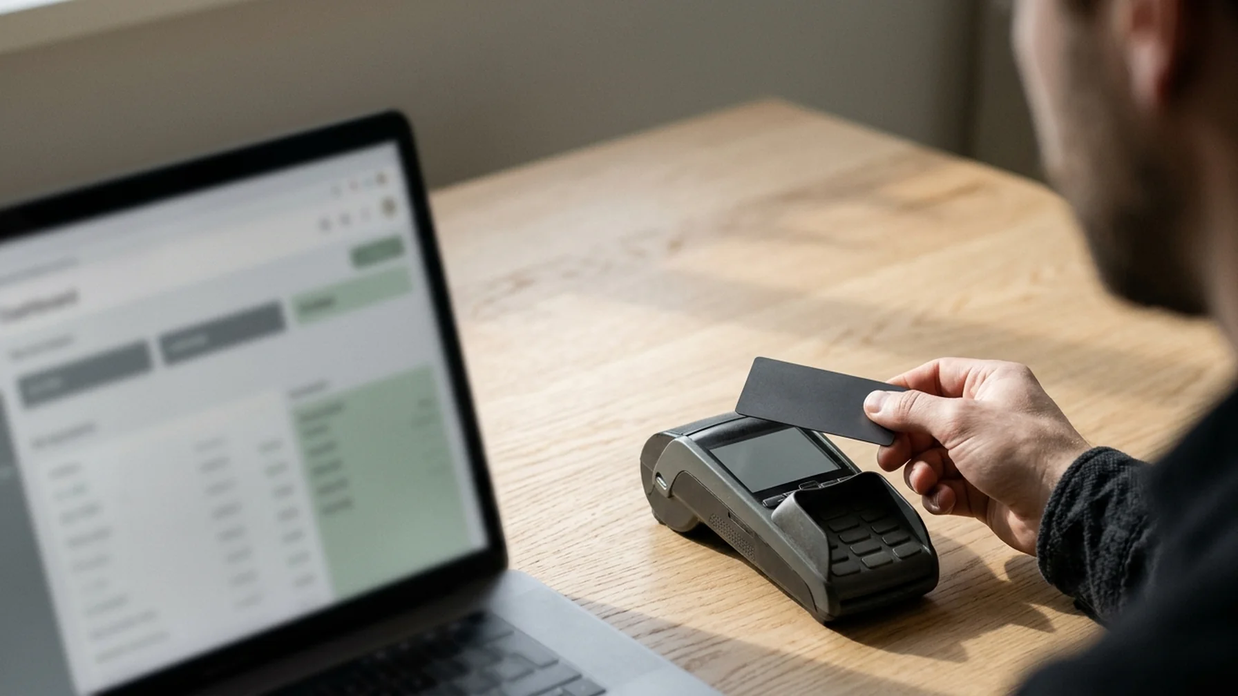 Close-up of a payment terminal on a light wood desk beside a laptop showing a billing dashboard, a card held in one hand, soft daylight from a side window